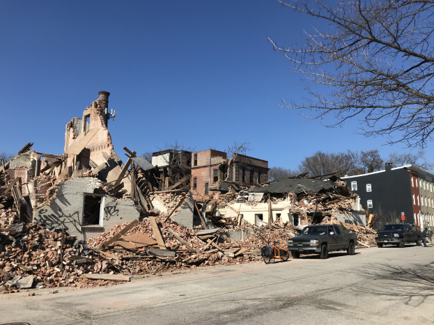 Ein eingestürztes Gebäude an einer Straßenkreuzung mit verstreuter Trümmern, Fahrzeugen, Passanten, Bäumen und einem klaren Himmel im Hintergrund.