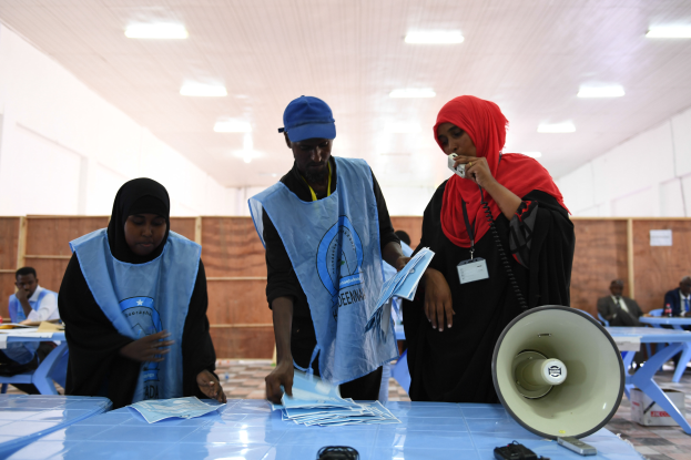 Eine Gruppe von Menschen steht um einen Tisch mit einem Megafon, Papieren und anderen Gegenständen, während andere im Hintergrund auf Stühlen sitzen oder stehen, unter einer Decke mit Lampen, was auf eine Wahllokalität hinweist.