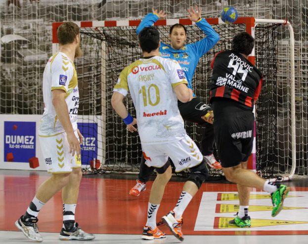 Eine Gruppe von Männern, die Handball auf einem Platz spielen, mit einem Ball in der Luft, einem Tor im Hintergrund und Texttafeln in einer Arena.