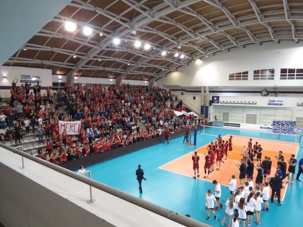Eine Gruppe von Menschen, die Volleyball in einer Turnhalle mit einem Netz spielt, Zuschauern auf Stühlen, Bannern und einer Wand mit Fenstern und Deckenleuchten.