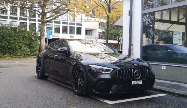Schwarzer Mercedes-AMG GT 63 S 4MATIC+ vor einem Gebäude mit Glasfenstern geparkt, mit Bäumen, Pflanzen, Schildern, Polen, Verkehrszeichen und anderen Gebäuden im Hintergrund.