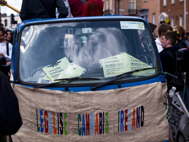 Eine Gruppe von Menschen, die um ein Auto mit einem "Legalise Cannabis Ireland"-Schild herumstehen, umgeben von Gebäuden unter einem klaren blauen Himmel, mit Papieren im Auto.