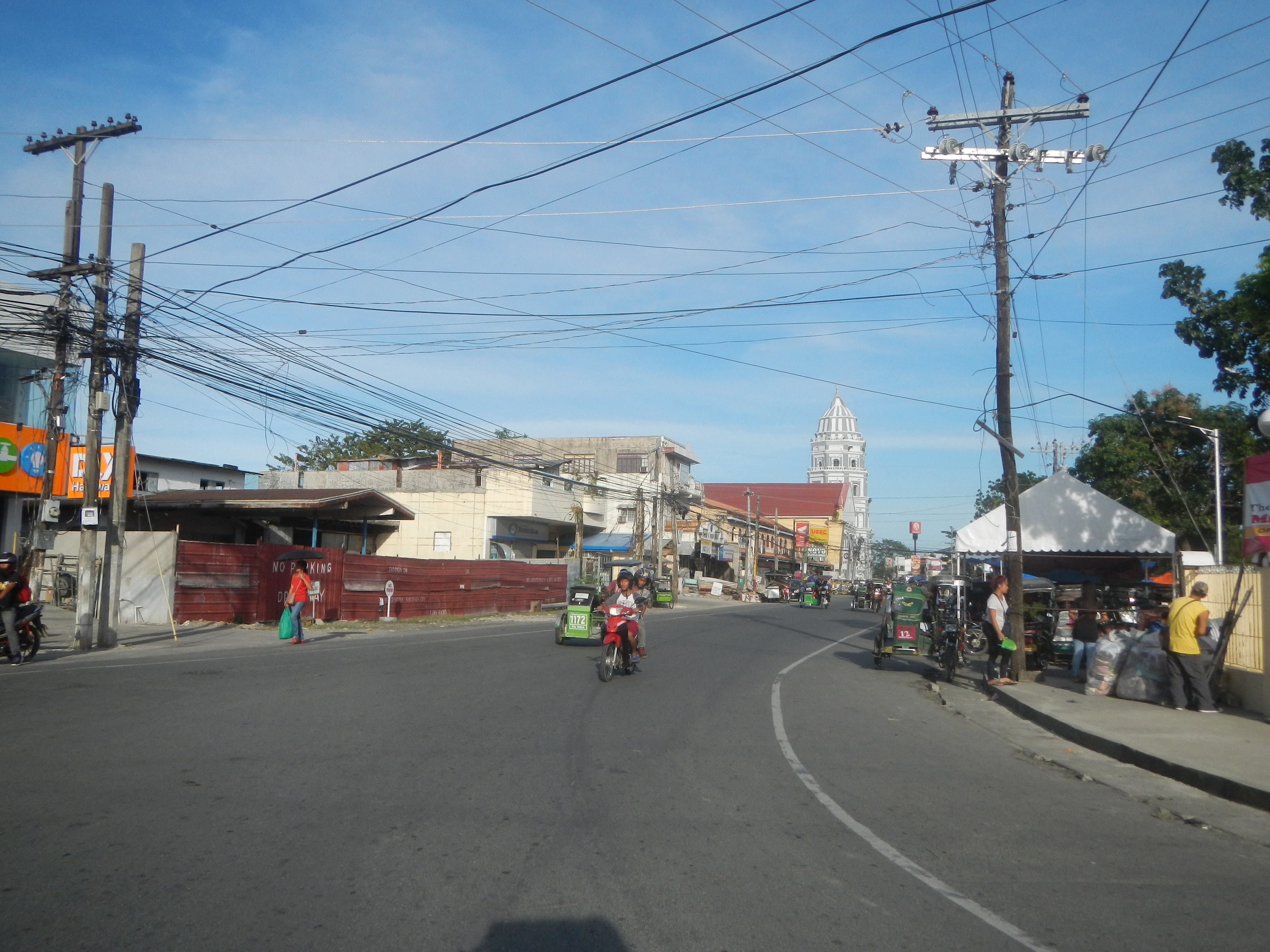 Eine Straße in einer Stadt mit Strommasten, Kabeln, Füßgängern, Motorradfahrern, Ständen, Bäumen, Gebäuden und einem klaren blauen Himmel.
