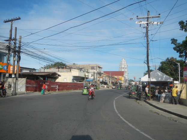Eine Straße in einer Stadt mit Strommasten, Kabeln, Füßgängern, Motorradfahrern, Ständen, Bäumen, Gebäuden und einem klaren blauen Himmel.