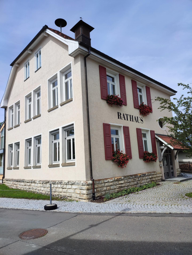 Weißes Gebäude mit roten Fensterläden, umgeben von Grün, mit einer Straße unten und Himmel im Hintergrund, identifiziert als das Rathaus Hotel in Baden-Württemberg.