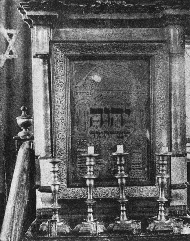 Schwarzes und weißes Foto eines Synagogeninneren mit einer Thorarolle an der Wand, Kerzenständern, einem Vorhang und anderen zeremoniellen Gegenständen, mit Text unten.