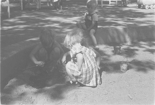 Drei Kinder beim Spielen im Sand, mit einem Jungen und einem Mödchen neben einer Schaufel, und anderen auf Stühlen mit einem Wagen rechts.