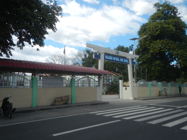 Einfahrt zu einer Schule mit einem vor der Tür geparkten Motorrad, mit einem Tor, einem Schuppen, einem Schild, Straßenmarkierungen, Laternen, Bäumen, einer Fahne und einem bewölkten Himmel.