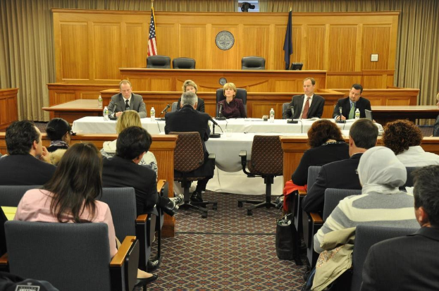 Ein Versammlungsraum mit sitzenden Menschen, zwei ausländischen Flaggen, einem orangen Schreibtisch, Wasserflaschen und einem Mikrofon.