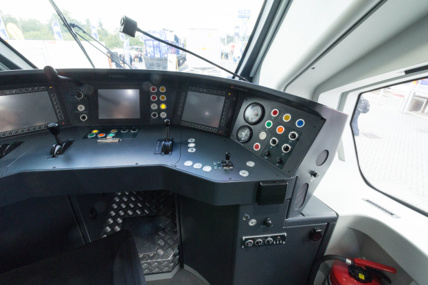 Cockpit eines Zuges mit einem Feuerlöscher im Vordergrund, das eine Glasscheibe mit Menschen, Fahrzeugen, Bäumen, Masten und anderen Gegenständen im Hintergrund zeigt.
