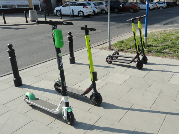 Drei Elektro-Scooter auf einem grasbewachsenen Straßenrand mit Gebäuden, Bäumen und Fahrzeugen im Hintergrund unter einem klaren blauen Himmel.