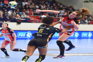 Eine Gruppe von Frauen beim Handballspielen auf einem Feld mit einem Ball in der Luft, während Zuschauer von Stühlen, Geländern und einer Wand im Hintergrund zusehen.