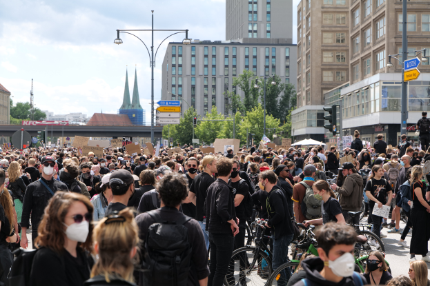 Eine große Gruppe von Menschen mit Masken geht eine Straße mit Bäumen, Laternen, Verkehrszeichen, Schildern und Gebäuden mit Fenstern entlang, einige halten Fahrräder, was auf eine Demonstration in Berlin hindeutet, mit einer bewölkten Himmel im Hintergrund.