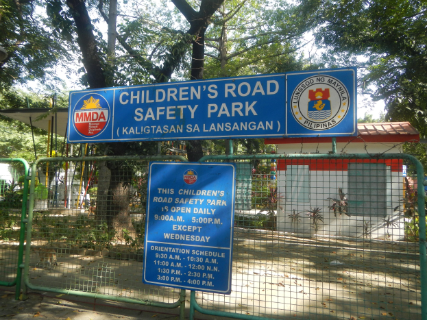 Metallzaun um einen Kinderverkehrssicherheitspark mit zwei blauen Informationsschildern, einem kleinen Haus mit Dach, Pflanzen, Bäumen und einem bewölkten Himmel.