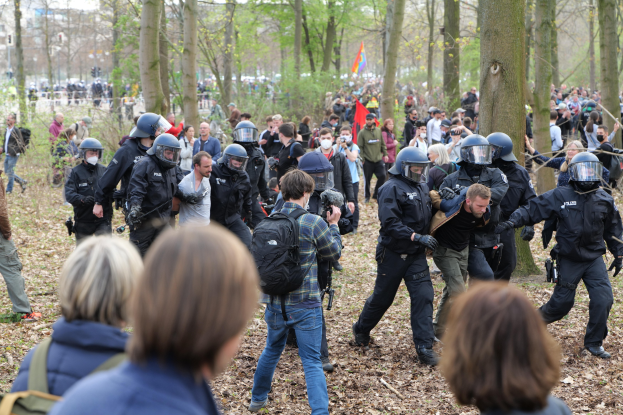 Polizei in Einsatzkleidung vor einer Menge mit Helmen und Taschen, mit Bäumen, einer Fahnenstange, abgefallenen Blättern und entfernten Gebäuden im Hintergrund.