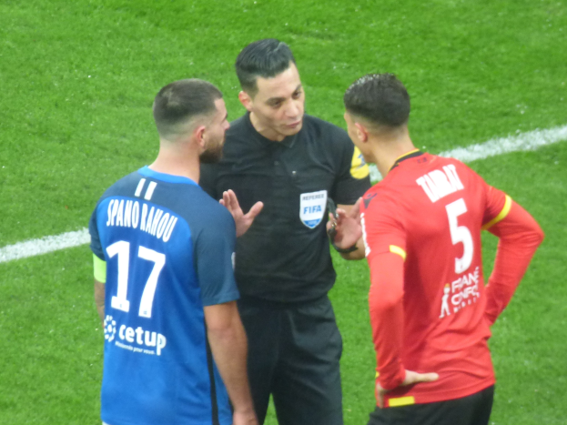 Zwei Männer in Fußballtrikots, einer in Schwarz, der andere in Blau und Rot, sprechen mit einem Schiedsrichter auf dem Feld, alle tragen Brillen.