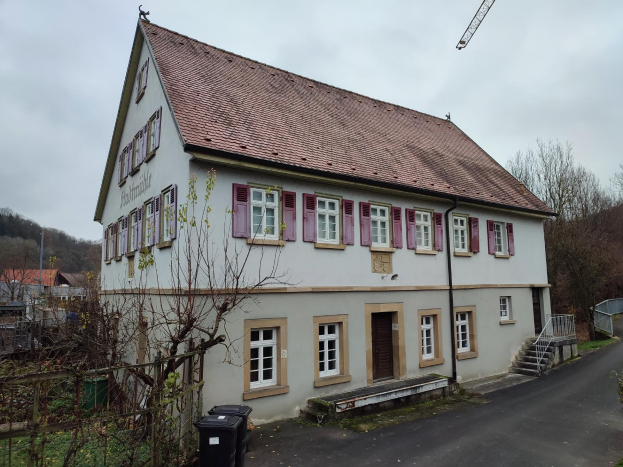 Großes weiße Haus mit roten Fensterläden, umgeben von Grün, Zaun und Außenobjekten, auf Hühllen unter einem klaren blauen Himmel, zum Verkauf in Baden-Württemberg.