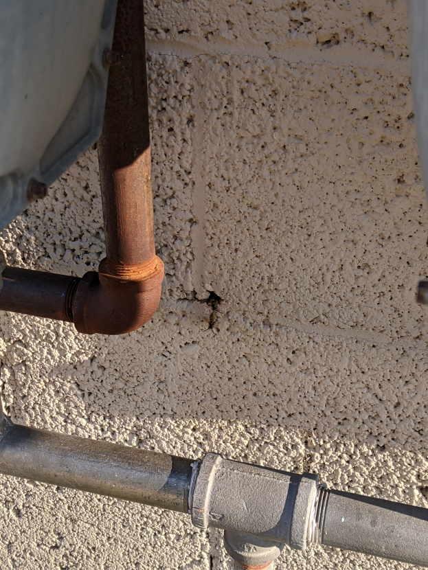 Nahaufnahme eines Metallrohrs, das an einem Metallwasserheizer an der Seite eines Gebäudes angeschlossen ist, mit einer Wand im Hintergrund.