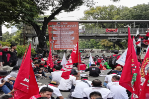 Eine große Gruppe von Menschen sitzt auf dem Boden vor einer Menge, einige halten Fahnen und tragen Mützen, mit einem Banner, Bäumen und einem klaren blauen Himmel im Hintergrund und einer Person mit Megafon auf der rechten Seite.
