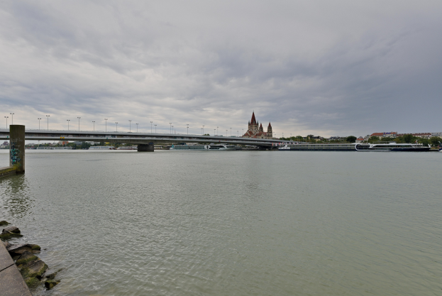 Eine steinerne Plattform auf der linken Seite bietet einen Blick auf die Donau in Budapest, Ungarn, mit einer Brücke, die das Wasser überspannt, Bäumen, Gebäuden und einem bewölkten Himmel im Hintergrund.