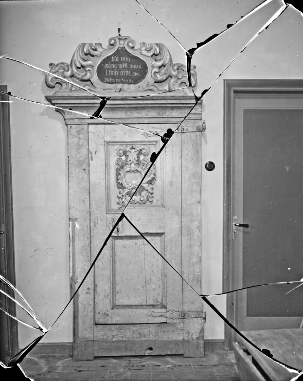 Ein Schwarz-Weiß-Foto einer zerbrochenen Tür in einem Raum mit verstreuten Glasscherben und einer Wand im Hintergrund.