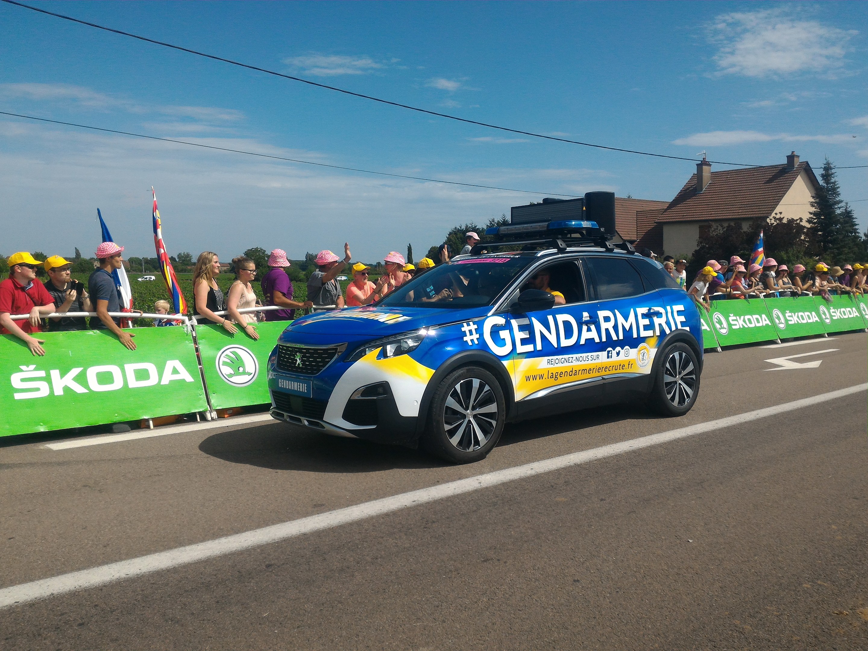 Polizeiauto fährt neben einer Menge, einige tragen Mützen und halten Fahnen, mit einem Banner auf dem Auto und Gebäuden, Bäumen und Wolken im Hintergrund; Text unten lautet "Gendarmerie de la Tour de France."