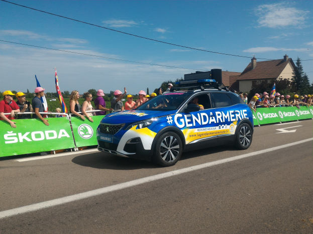 Polizeiauto fährt neben einer Menge, einige tragen Mützen und halten Fahnen, mit einem Banner auf dem Auto und Gebäuden, Bäumen und Wolken im Hintergrund; Text unten lautet "Gendarmerie de la Tour de France."