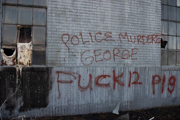 Graffiti auf einer Gebäudewand mit der Aufschrift 'Polizeimord George Fuck 12 P19' mit Fenstern und verstreuten Gegenständen davor.
