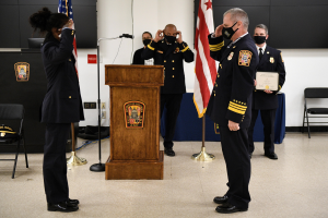 Gruppe uniformierter Personen bei einer Vereidigung eines neuen Feuerwehrchefs, der ein Zertifikat hält, mit Fahnen, Podium und maskierten Teilnehmern im Hintergrund.