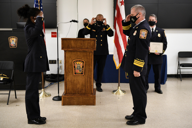 Gruppe uniformierter Personen bei einer Vereidigung eines neuen Feuerwehrchefs, der ein Zertifikat hält, mit Fahnen, Podium und maskierten Teilnehmern im Hintergrund.