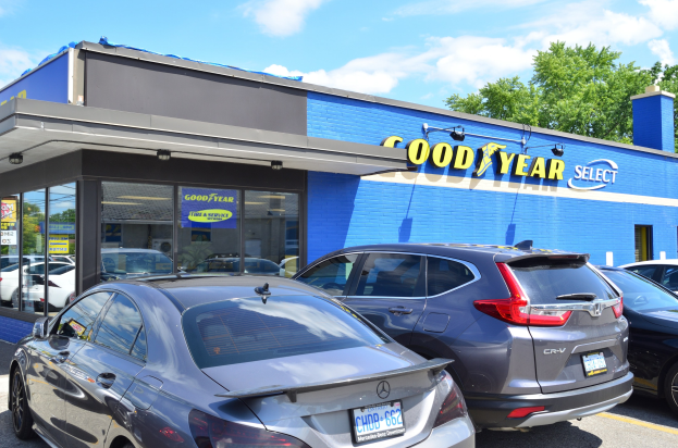 Ein Goodyear Select Autohaus mit parkenden Fahrzeugen vorne, Text auf dem Gebäude, Bäumen im Hintergrund und einem bewölkten Himmel.