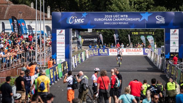 Eine Gruppe von Radfahrern fährt unter einem Bogen mit der Aufschrift "2019 UEC MTB Europameisterschaften" eine Straße hinunter, während Zuschauer auf beiden Seiten stehen und Bäume, Gebäude und ein klarer blauer Himmel im Hintergrund zu sehen sind.