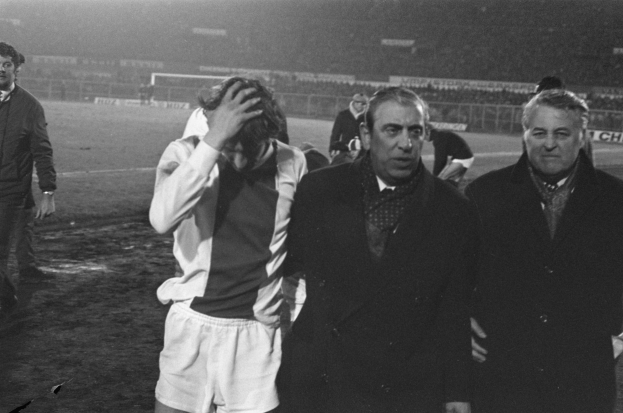 Schwarzes und weißes Bild von Männern in Fußballtrikots, die auf einem Feld mit einem Stadion im Hintergrund stehen und enttäuscht nach einem Matchsieg aussehen.