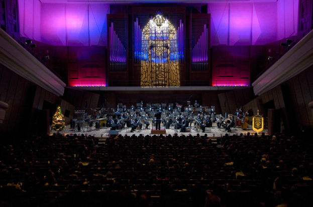 Großer Saal mit Publikum bei einem Konzert, Musiker auf der Bühne und sitzend, Weihnachtsbaum im Hintergrund, Wände mit Lichtern und Schmuck dekoriert.