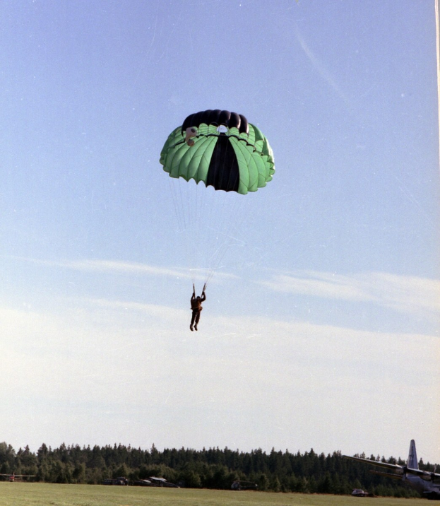 Eine Person beim Paragliding in der Luft mit einem Fallschirm, umgeben von Gras und Bäumen, mit einem Flugzeug auf der rechten Seite und Wolken am Himmel.