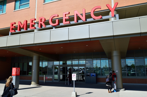 Gruppe von Menschen geht auf ein Notaufnahmegebäude mit Glaswänden, Säulen und einem Namensschild zu, vorbei an Schildern und einem Müllcontainer.