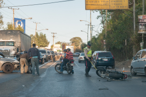 Gruppe von Menschen um ein verunglücktes Motorrad auf dem Seitenstreifen mit mehreren Fahrzeugen, darunter ein Lastwagen, im Hintergrund und Bäumen, Masten, Lichtern, Schildern und Himmel im Bild.