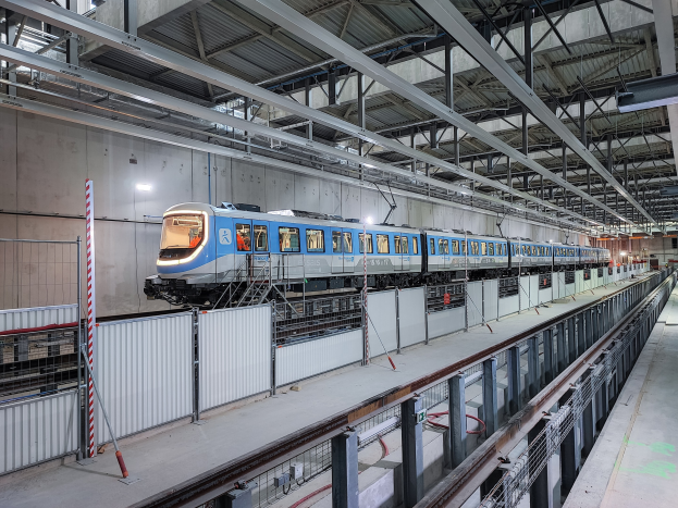 Blauer und weißer Zug fährt durch eine große Innenstation mit einer Plattform auf der rechten Seite und einer Wand im Hintergrund, unter einer Decke mit Stangen und Lampen.