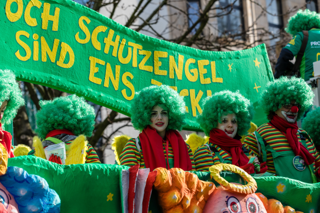 Eine Gruppe von Clowns in bunten Kostümen marschiert durch die Straße, hält ein Banner mit Text, mit Bäumen und einem Gebäude im Hintergrund.