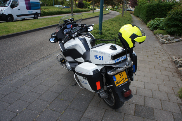 Ein Polizeimotorrad mit einem Helm darauf auf der Seite einer Straße geparkt, mit Bäumen, Pfählen, Gebäuden, Pflanzen, Gras, Steinen und anderen Fahrzeugen im Hintergrund.