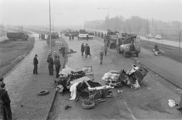 Schwarz-weiß-Szene eines Autounfalls am Straßenrand mit mehreren Fahrzeugen, Einsatzkräften und Schaulustigen, Laternenpfähle, Bäume und Himmel im Hintergrund.