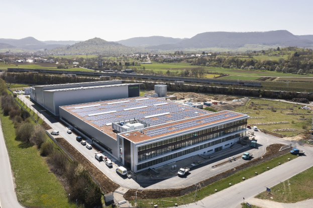 Luftaufnahme eines großen Gebäudes mit Solarmodulen auf dem Dach, umgeben von Fahrzeugen, Bäumen, Gras und einem klaren blauen Himmel im Hintergrund.