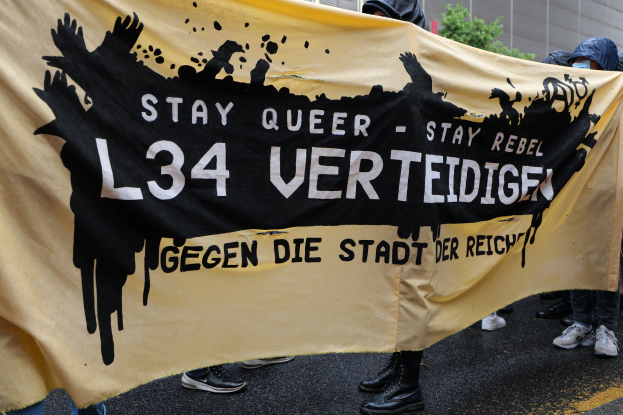 Gruppe von Menschen mit einer gelben Fahne, auf der "Stay Queer - Stay Rebel L34 Verteidige" steht, während einer Demonstration gegen die L34-Bewegung, mit einem Gebäude, einem Baum und dem Himmel im Hintergrund.