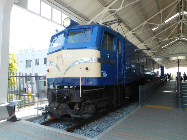 Blauer und weißer Zug fährt auf Schienen neben einem Bahnsteig mit einigen Menschen, Treppe auf der rechten Seite, Geländer auf der linken Seite und Hintergrund mit einem Eisenzaun, Bäumen, einem Gebäude und Himmel.