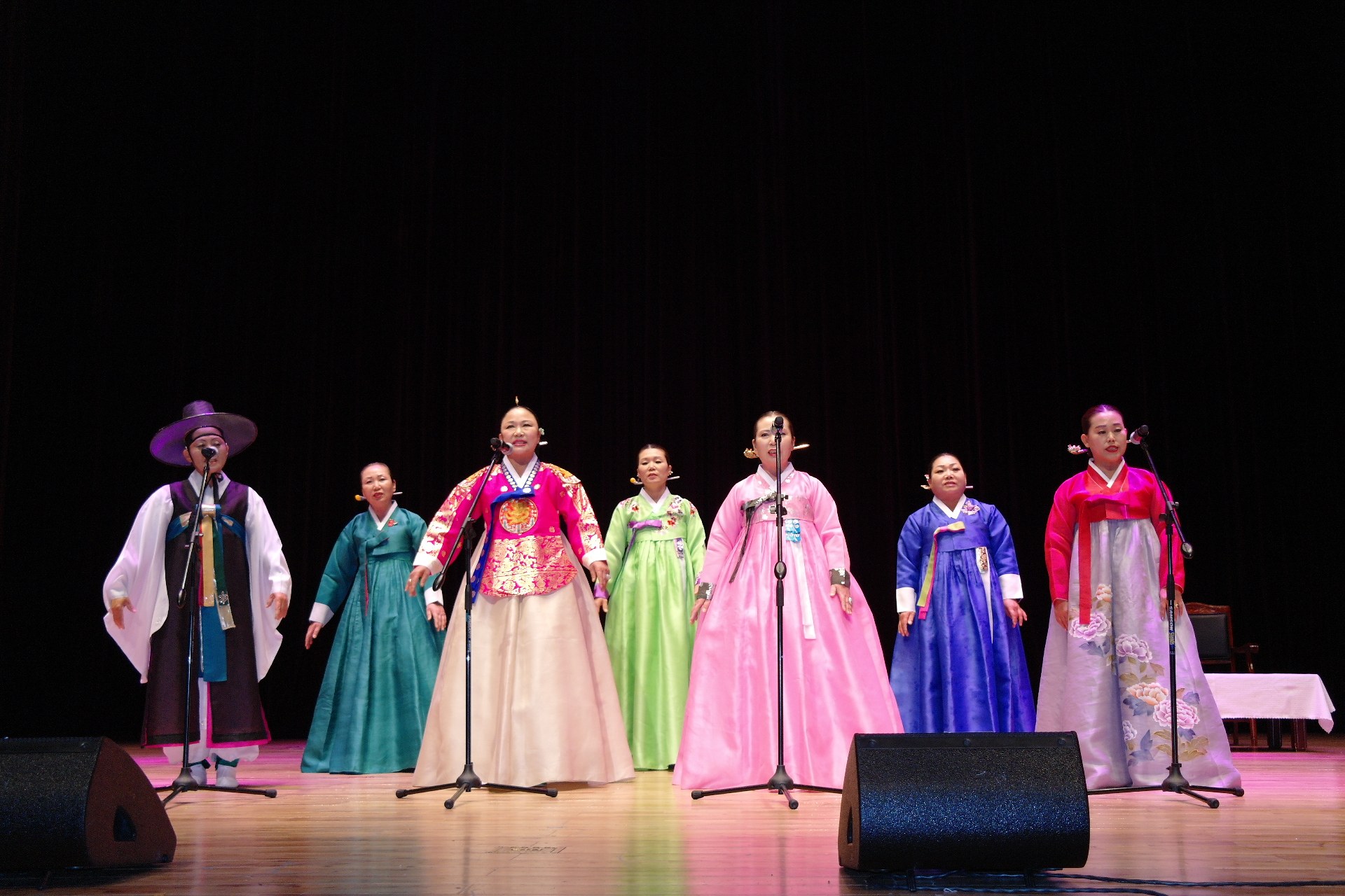 Eine Gruppe von Menschen in bunten Kleidern, die in Mikrofone singen, auf einer Bühne mit Lautsprechern und einem gedeckten Tisch, vor einem dunklen Hintergrund.