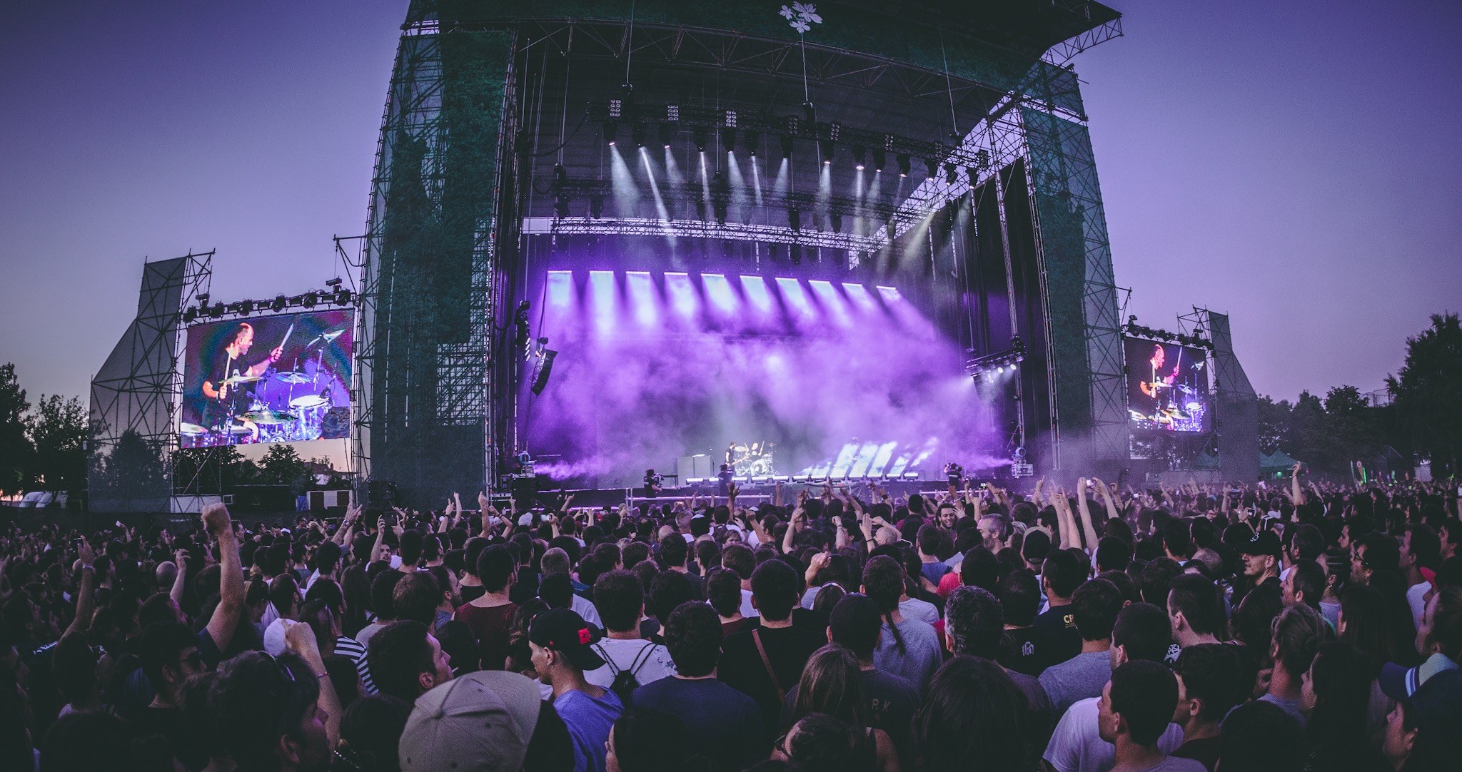 Eine große Menschenmenge versammelt sich auf einem Musikfestival mit einer Bühne, auf der Musiker spielen, die von Scheinwerfern beleuchtet und von Bildschirmen umgeben sind, vor einer Kulisse aus einem Gebäude, Bäumen und einem klaren blauen Himmel.