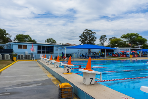 Großer Swimmingpool mit schwimmenden Menschen, umgeben von Bahnmarkierungen, Verkehrskegeln, Stühlen, Sonnenschirmen, einem Gebäude mit Fenstern, einer Flagge, Bäumen und einem bewölkten Himmel im Hintergrund.