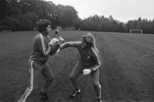 Zwei Männer in schwarzer und weißer Fußballuniform mit Handschuhen geraten auf einem Feld mit Bäumen und einer Hütte im Hintergrund in eine körperliche Auseinandersetzung.