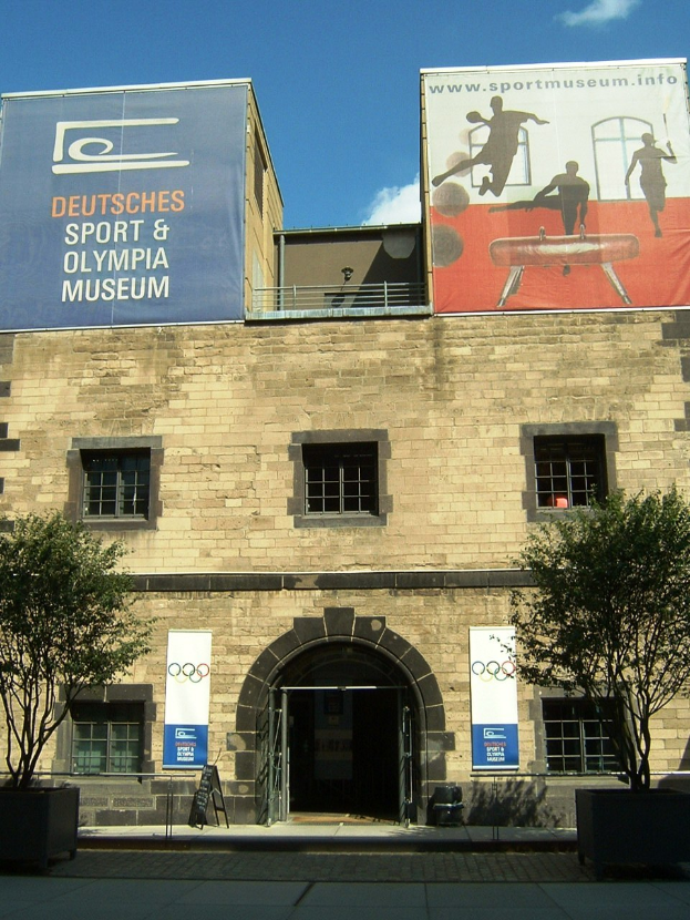 Das Deutsche Sport & Olympia Museum in Berlin, Deutschland, ein Gebäude mit Fenstern und einer Tür, umgeben von Bäumen und Bannern mit Text und Bildern, unter einem bewölkten Himmel.