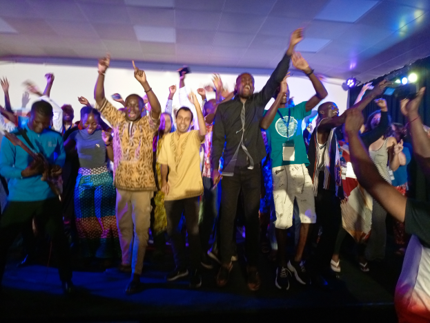 Eine Gruppe von Menschen, die auf einer Bühne tanzen und die Hände erhoben haben, beleuchtet von Scheinwerfern auf der rechten Seite und einem Bildschirm im Hintergrund, unter einer Decke mit Scheinwerfern.